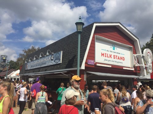 A busy milk stand with a red barn-style building, people lining up, and a sign reading "All You Can Drink Milk Stand."