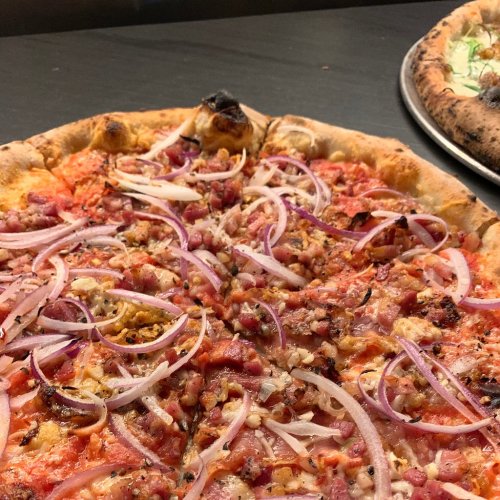 Close-up of a pizza topped with red onions, meats, and a golden crust, with another pizza in the background.