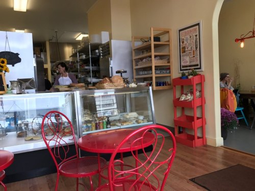Cozy café interior with red chairs, a display case of baked goods, and a barista preparing food in the background.