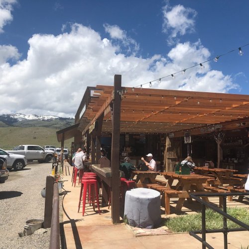 A rustic outdoor dining area with wooden tables and chairs, surrounded by mountains and blue skies.
