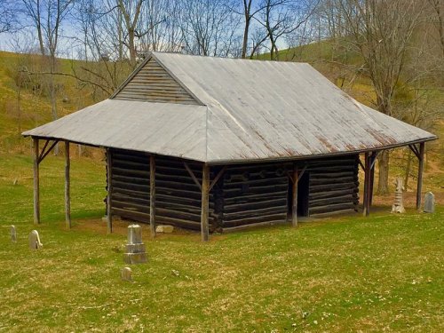 A rustic log cabin with a metal roof, surrounded by grass and trees, near a small graveyard.