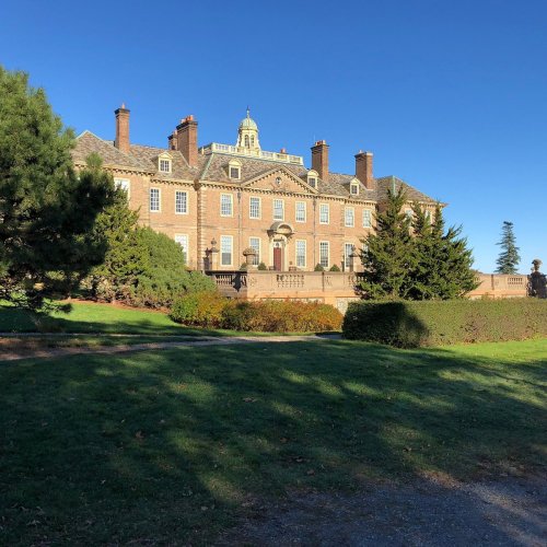 A large historic mansion surrounded by greenery and manicured lawns under a clear blue sky.