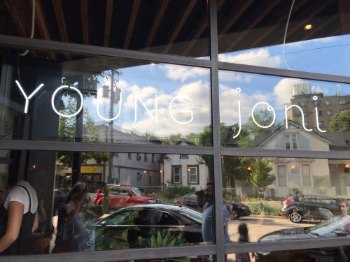 Sign for "YOUNG joni" on a restaurant window, reflecting the street and people outside.