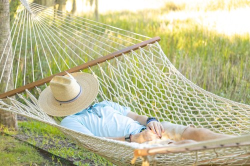 A person wearing a wide-brimmed hat relaxes in a hammock surrounded by greenery.