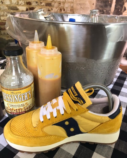 A yellow sneaker rests on a checkered tablecloth next to a bottle of sauce and a metal bucket filled with ice.
