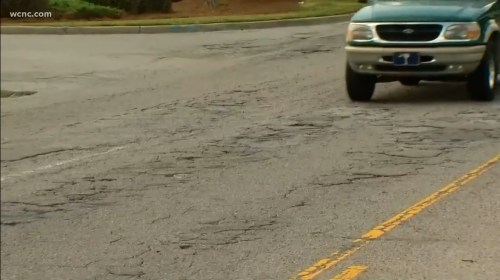 A vehicle drives on a road with visible cracks and potholes, indicating poor road conditions.