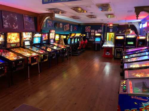 A vibrant arcade filled with pinball machines and arcade games, featuring colorful lights and a wooden floor.