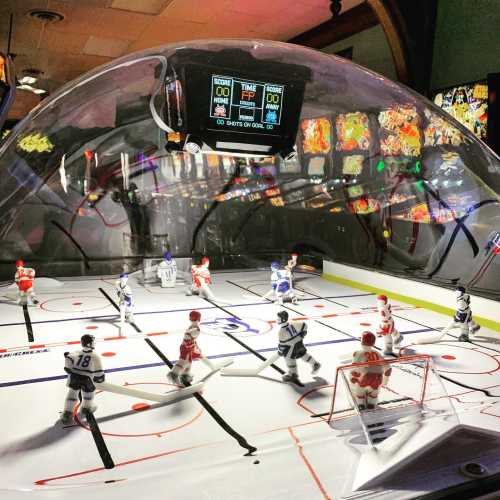 A tabletop hockey game with players in blue and red jerseys, under a dome, with a scoreboard in the background.