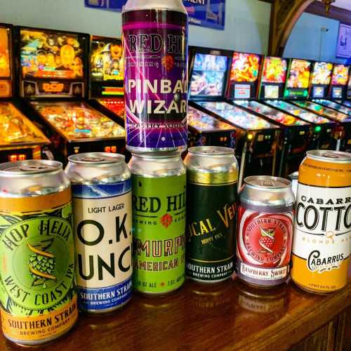 A collection of craft beer cans in front of colorful pinball machines in a lively arcade setting.