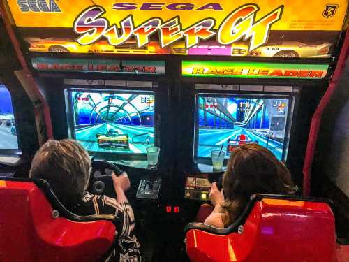 Two players enjoying a Sega Super GT arcade racing game, focused on their screens and steering wheels.