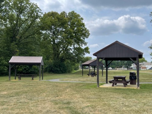 Three wooden pavilions with picnic tables in a grassy park under a partly cloudy sky.