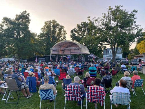 A large crowd sits on lawn chairs, enjoying a live music performance on an outdoor stage surrounded by trees.