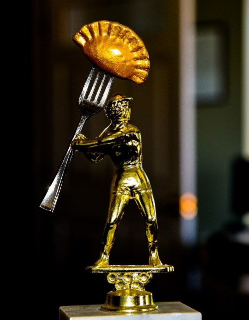 A golden trophy figure holding a fork with a golden empanada on top, set against a blurred background.