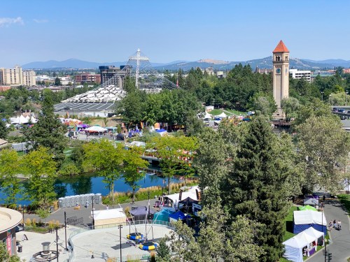 A vibrant park scene with a clock tower, trees, a pond, and festival tents under a clear blue sky.
