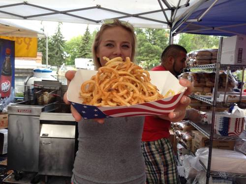A person holds a tray of curly fries under a tent at a food festival, with a grill and food items in the background.