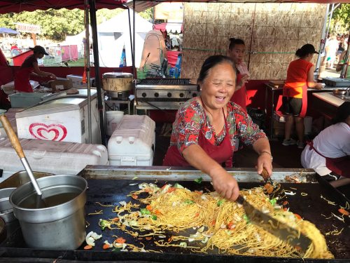 A smiling woman cooks noodles at a vibrant outdoor food stall, surrounded by colorful tents and people.