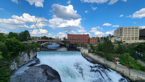 A scenic view of a waterfall with a city skyline, blue sky, and fluffy clouds in the background.