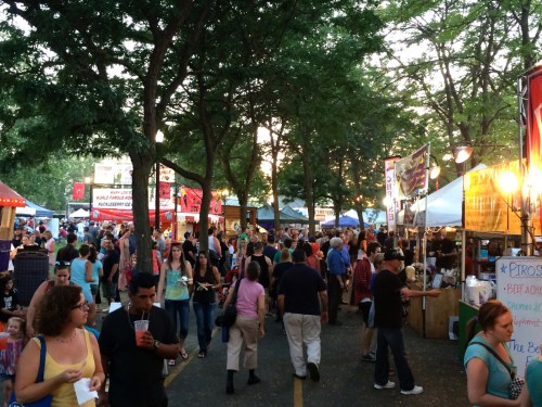 A bustling outdoor market with food stalls and crowds under trees, creating a lively atmosphere.