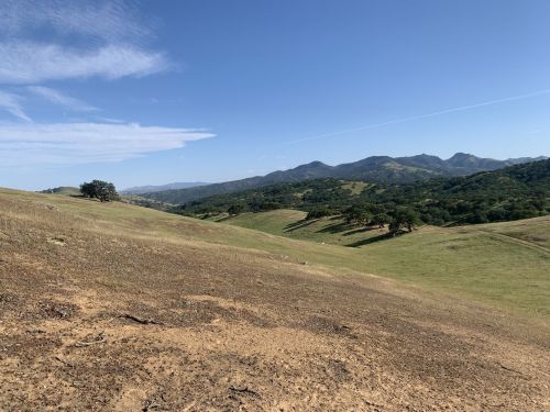 A scenic landscape featuring rolling hills, green valleys, and distant mountains under a clear blue sky.