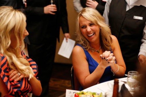 Two women at a social gathering, one in a blue dress laughing and enjoying the moment, with others in the background.