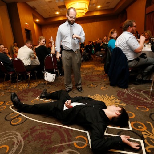 A man in a suit lies on the floor while another man stands nearby, speaking to a crowd in a banquet hall.