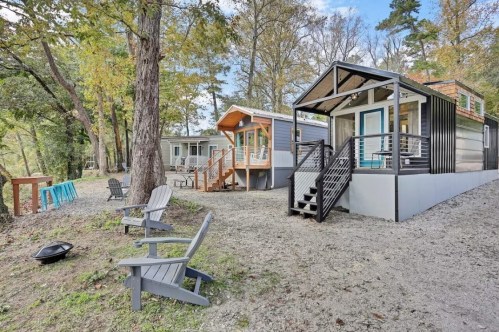 Two modern tiny houses with porches, surrounded by trees and gravel, with outdoor seating and a fire pit nearby.