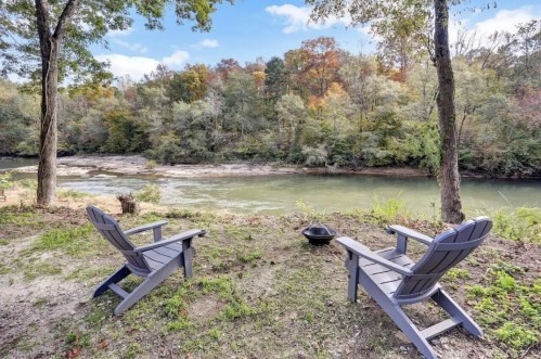 Two gray Adirondack chairs facing a serene river surrounded by trees with autumn foliage.