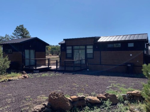 Two modern tiny homes with wooden exteriors, surrounded by gravel and greenery under a clear blue sky.