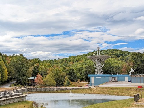 A satellite dish stands near a pond, surrounded by trees and a blue sky with clouds in a scenic landscape.