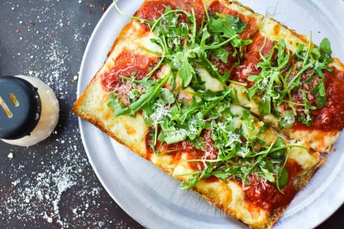 A rectangular pizza topped with marinara sauce, cheese, and fresh arugula, served on a metal plate with grated cheese nearby.