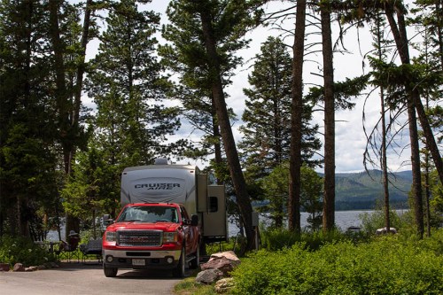 A red truck parked beside a camper in a forested area near a lake, surrounded by tall trees and mountains in the background.