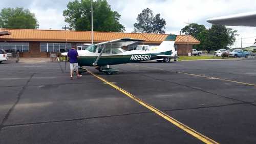 A small green and white airplane parked on a tarmac, with a person standing nearby and a building in the background.