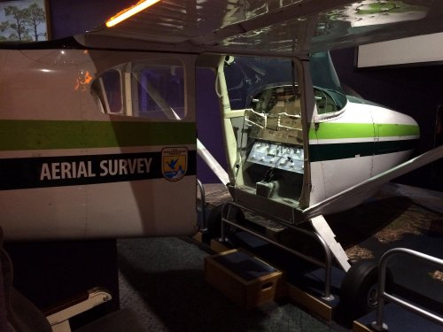 A small aircraft with an open cockpit, labeled "AERIAL SURVEY," displayed in a museum setting.