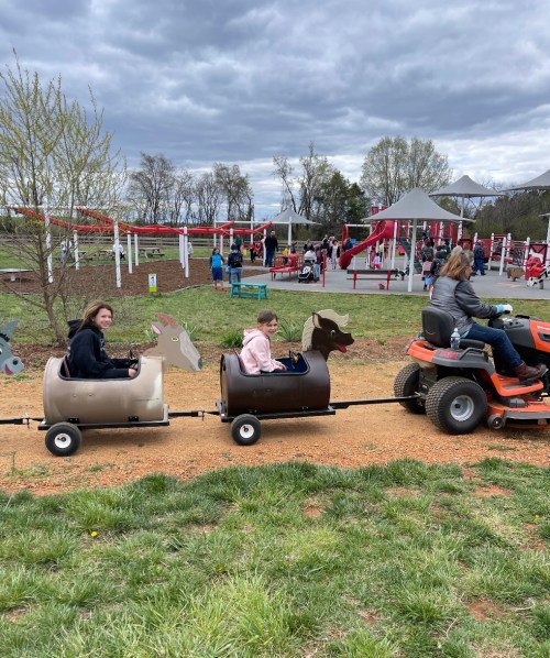 Two children ride in barrel-shaped carts resembling animals, pulled by a lawn tractor at a playground.