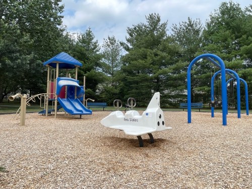 A playground featuring a slide, swings, and a space shuttle-shaped play structure on a wood chip surface.