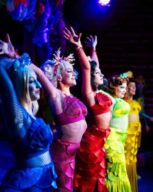 A group of performers in colorful, layered costumes pose with arms raised on stage, illuminated by vibrant lights.