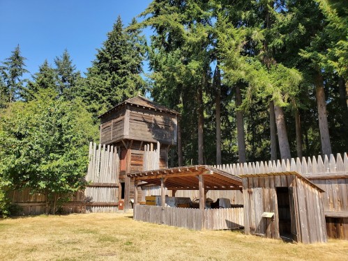A wooden fort with a lookout tower surrounded by tall trees and a grassy area.