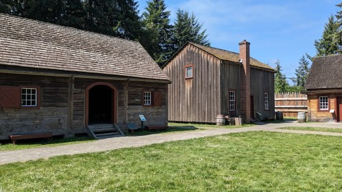 Two historic wooden buildings with sloped roofs, surrounded by green grass and trees under a clear blue sky.