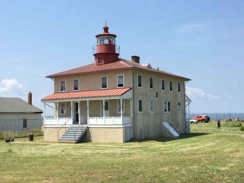A historic lighthouse with a red roof and lantern, surrounded by green grass and a clear blue sky.