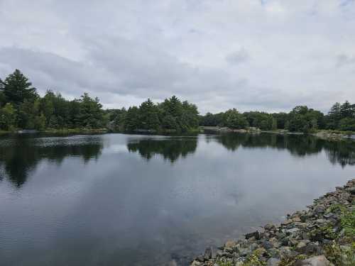 A calm lake surrounded by trees, with a rocky shoreline and cloudy skies reflecting on the water's surface.