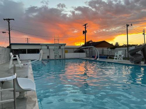 A swimming pool at sunset, with vibrant orange and purple skies, surrounded by a fence and poolside chairs.
