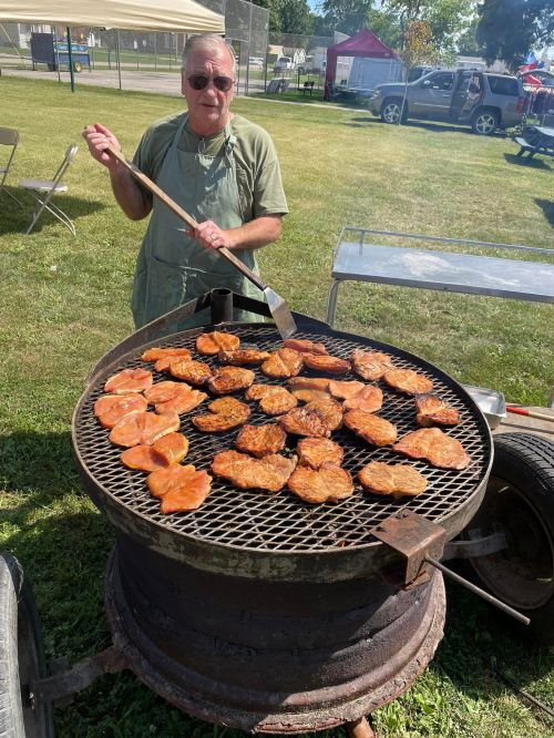 A man in an apron grills multiple pieces of meat on a large barbecue in a grassy area.