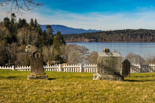 A serene graveyard with old tombstones overlooking a calm lake and distant mountains under a clear blue sky.