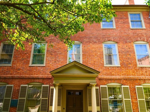 Historic brick building with large windows and a central entrance, framed by green leaves from a nearby tree.