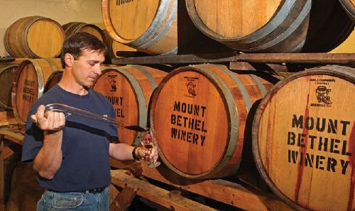 A man samples wine from a barrel at Mount Bethel Winery, surrounded by stacked wooden barrels.