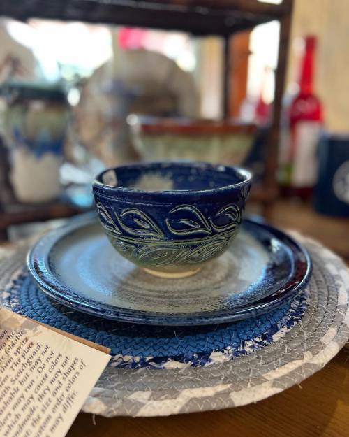 A decorative ceramic bowl on a plate, displayed on a woven mat with other pottery in the background.