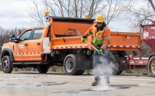 A worker in a yellow hard hat and orange vest repairs a road with a tool, while an orange truck is parked nearby.