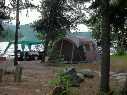 A tent set up by a lake, surrounded by trees, with a green canopy nearby and rocky ground.