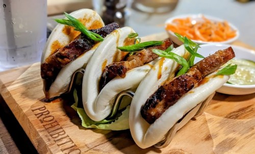 Three bao buns filled with grilled meat, garnished with green onions, served on a wooden platter with dipping sauce.