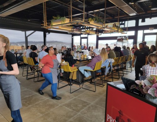 A busy restaurant interior with diners at a large table and servers attending to guests, featuring large windows and plants.